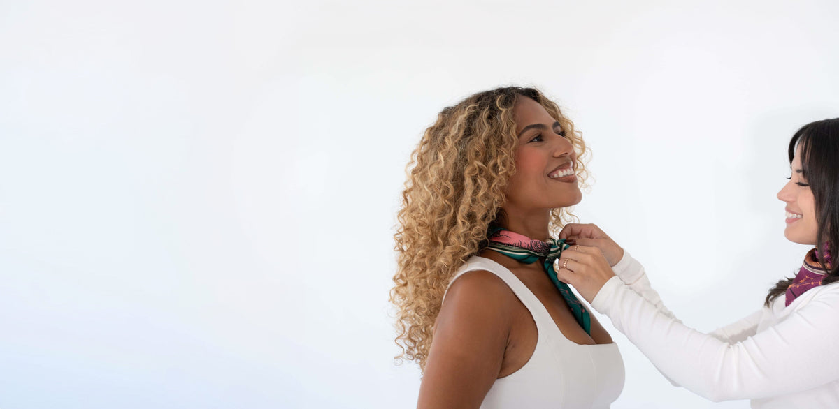 A woman with curly blonde hair wearing a white tank top smiles while another woman adjusts a green and pink neck scarf on her. The background is a clean, bright white studio setting.