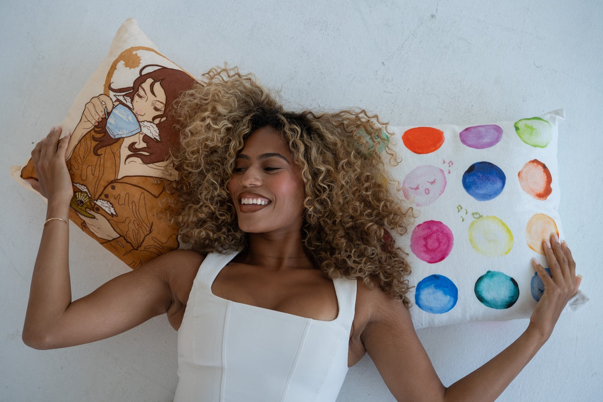A smiling woman with curly hair lies on a white wall between two decorative pillows. One pillow features a warm-toned illustrated design, and the other has a white background with colorful watercolor-style dots in pink, blue, orange, green, and purple.