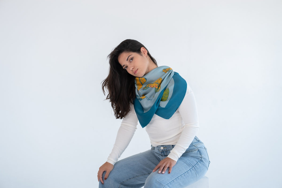 A woman wearing a white long-sleeve top and light blue jeans sits against a plain white studio background, draped with a teal and light blue scarf featuring a yellow and green abstract pattern.