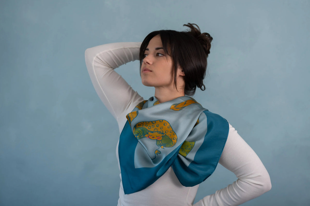 A woman wearing a white long-sleeve top and a blue silk scarf with a yellow and green floral pattern, posing against a textured blue background.