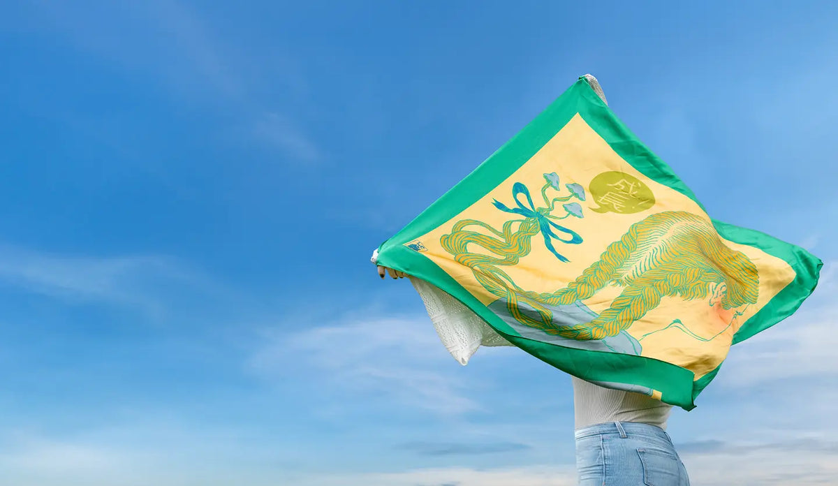 A square scarf with a green border and a yellow center featuring a green dragon design, held up by a person against a bright blue sky.