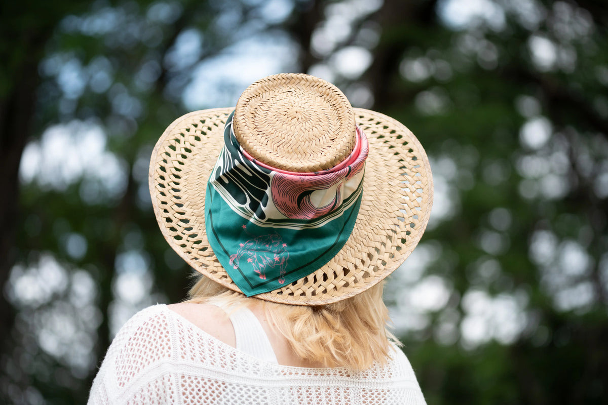 A woman viewed from behind wearing a natural straw wide-brim hat with a teal, pink, and black patterned silk bandana tied around the crown, set against a blurred green outdoor background.